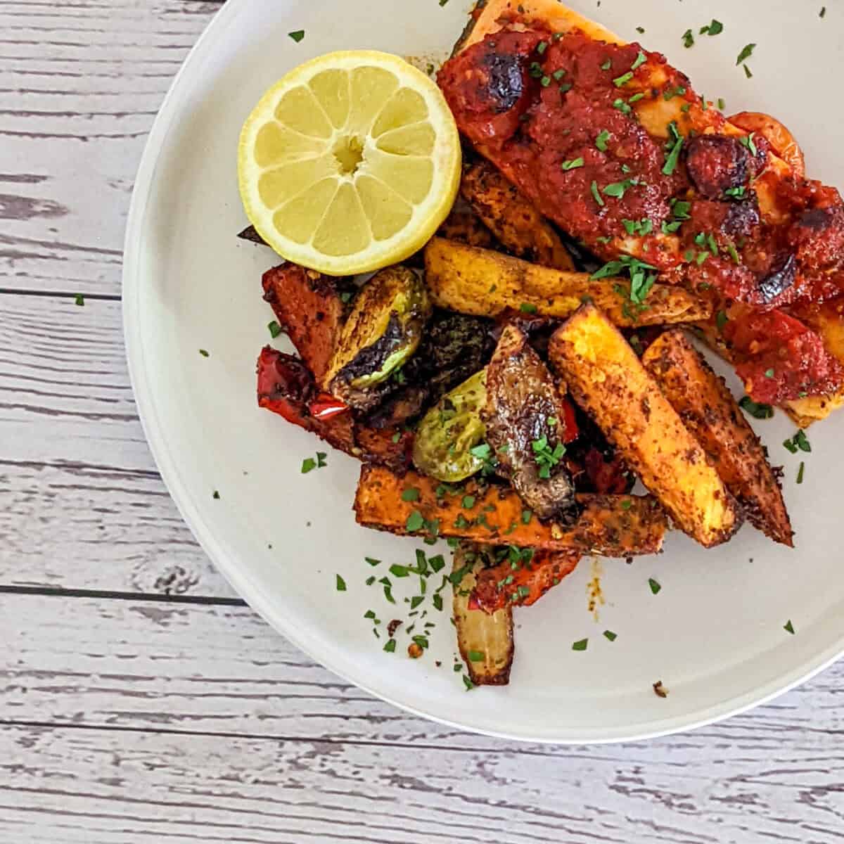 Roasted harissa salmon with paprika potatoes and brussel sprouts with half a lemon on a round white plate with parsley sprinkled on top.